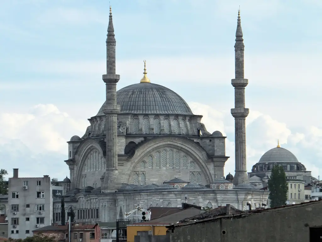 Nuruosmaniye Camii mahalle çatılarının üzerinden; kurşun kaplı tek büyük kubbe, iki ince minare, sağda külliyenin küçük kubbesi ve beyaz bulutlu mavi gökyüzü.