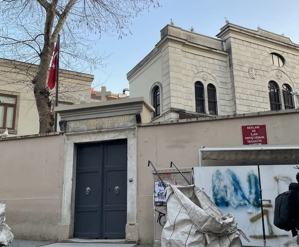 Surp Takavor Kilisesi'nin avlu kapısı; lacivert metal kapı, taş cephe, yuvarlak pencereler ve kapının önünde afişler.