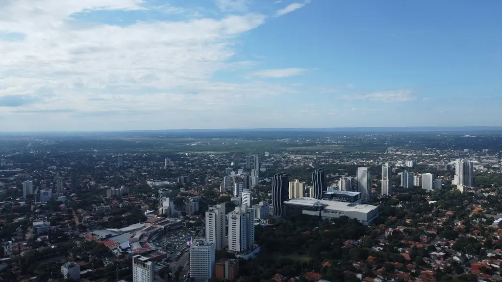 Paraguay'ın başkenti Asunción'un kuş bakışı görünümü; modern gökdelenler, kırmızı çatılı mahalleler ve ufka uzanan yeşil ovalar, açık mavi gökyüzü.