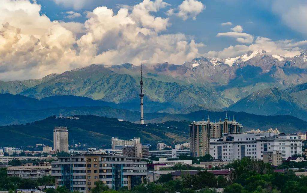Almatı'da yukarıdan görünen şehir; ağaçların arasına dağılmış apartmanlar, tepedeki televizyon kulesi ve arkada boydan boya uzanan karlı dağ sırası.