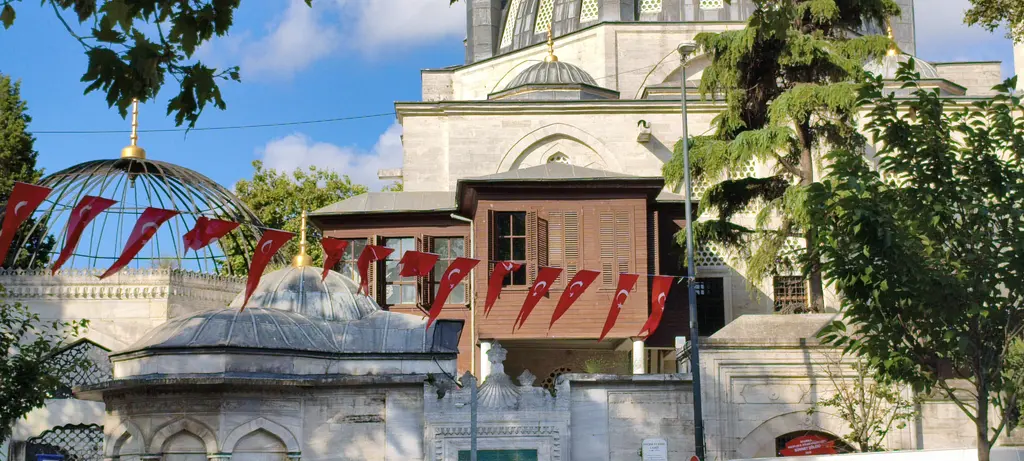 Yeni Valide Camii'nde hünkâr kasrı; kahverengi ahşap köşk, önde şebekeli türbe ve kırmızı bayraklar.