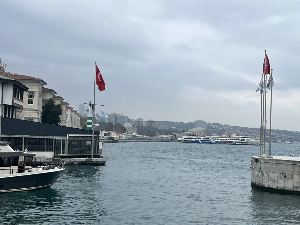 Galataport'ta iskelenin ucu; sağda Türk bayrağı, solda bağlı tekneler ve kıyı binaları, arkada Boğaz'ın suyu.