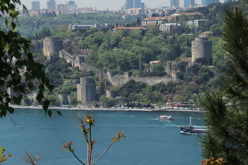 Sarıyer'de Rumeli Hisarı karşı kıyıdan; yeşil yamaca tırmanan kuleler ve surlar, mavi Boğaz ve önde çam dalları.