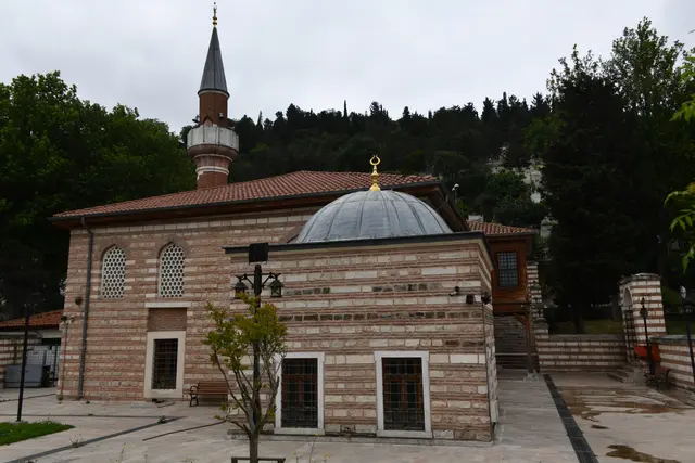Eyüp'te Şah Sultan Camii; tuğla ve taş almaşık duvarlar, kiremit çatı, kubbeli küçük türbe, minare ve arkada yeşil yamaç.