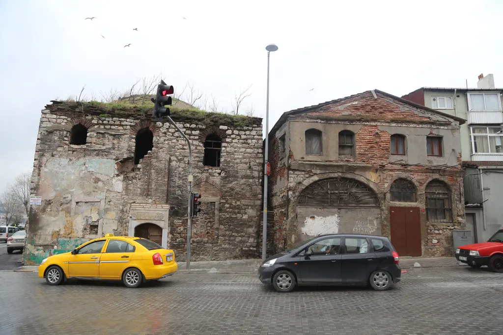 Ayakapı Hamamı ve yanındaki taş ev; harap duvarlar, kemerli pencereler, önde sarı taksi ve siyah araba.