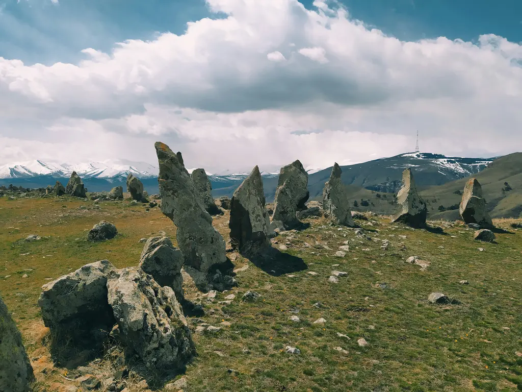 Sisian yakınında geniş bir bozkır platosunda dikili duran, bazılarının üstünde delik bulunan iri taş bloklar; arkada karla kaplı dağ sırası ve bulutlu gökyüzü.