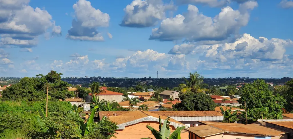 Kamerun'da bir kasabanın kiremit ve teneke çatıları; muz ve palmiye ağaçları arasına yayılan yerleşim ve ufuktaki alçak tepeler, bulutlu mavi gökyüzü.