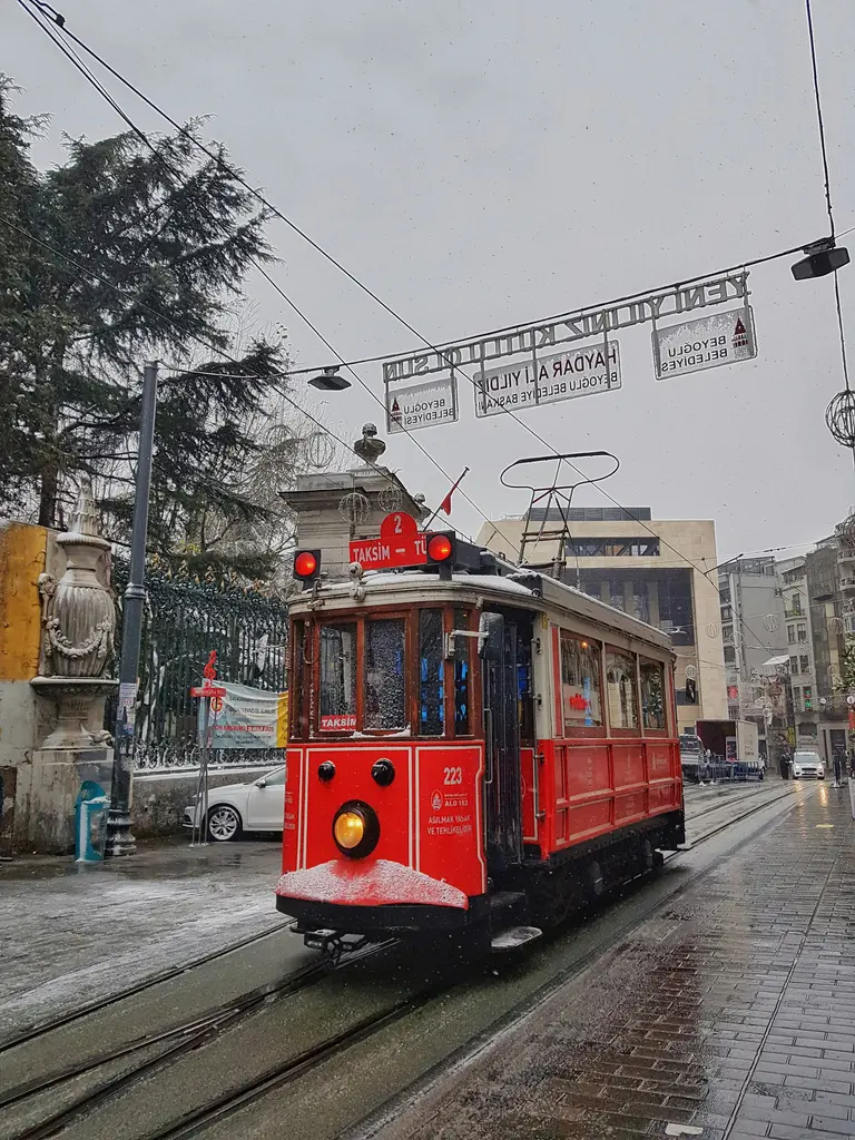 Beyoğlu'nda karlı bir günde kırmızı nostaljik tramvay; ıslak zemin, arkada karla örtülü ağaçlar ve üstte uzanan tramvay telleri.