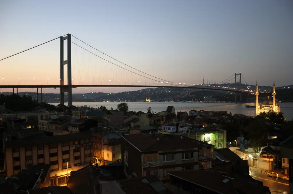 Akşam alacasında Ortaköy'e yukarıdan bakış; çatılar, ışıkları yanan evler, sağda aydınlatılmış cami ve üstte asma köprü.