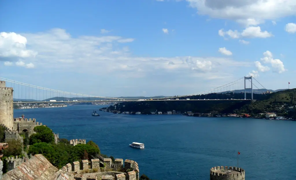 Rumeli Hisarı'ndan Boğaz; önde mazgallı sur ve kule, mavi deniz, tur teknesi ve uzakta asma köprü.
