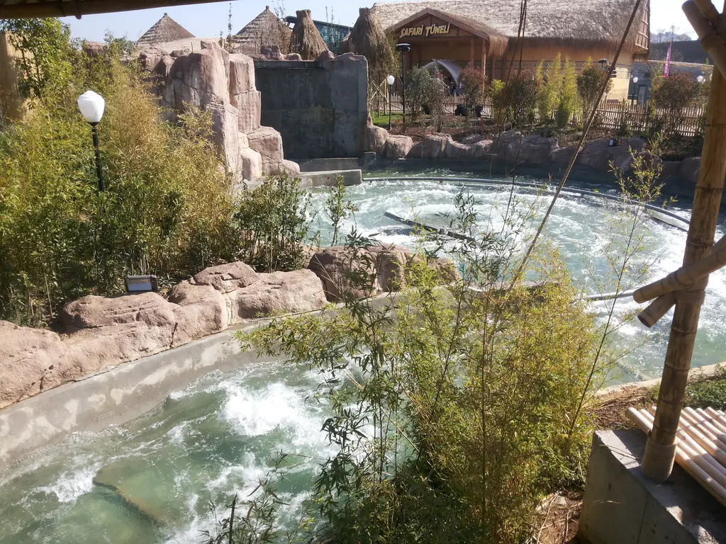 Vialand'de yapay kayalar arasında köpüklü sularıyla akan nehir parkuru; kıyıda bitkiler, ahşap yapılar ve bir aydınlatma direği.
