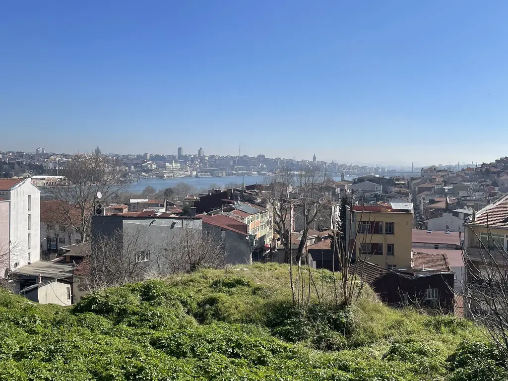 Balat'ın yamacından Haliç'e bakış; yeşil bir arsa, kiremit çatılar, eski binalar ve uzakta Haliç ile karşı kıyı.