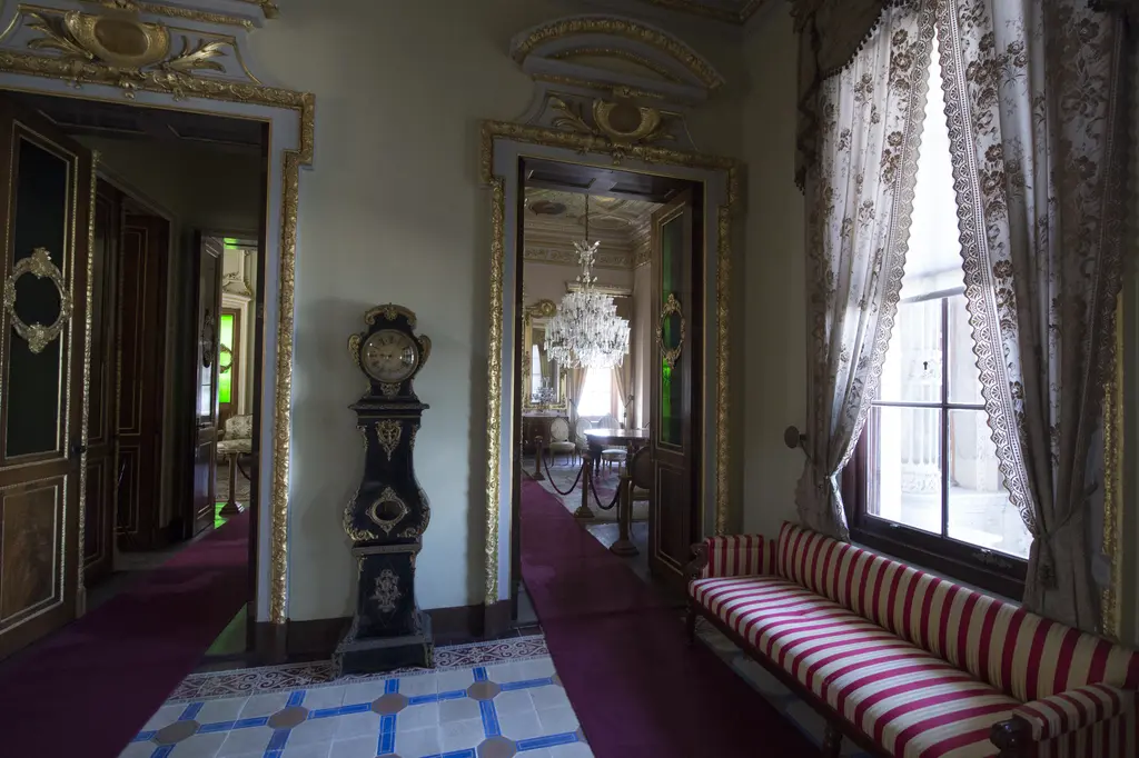 Küçüksu Kasrı'nda sofa; kapı arasında ayaklı bir saat, pencere önünde kırmızı-beyaz çizgili sedir ve renkli karo döşeme.