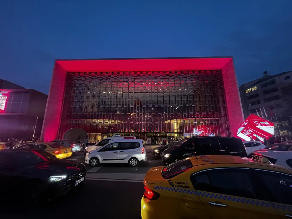 Atatürk Kültür Merkezi gece; pembe ve kırmızı ışıkla aydınlatılmış cephe, önde caddede ilerleyen taksi ve otomobiller.