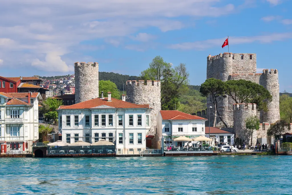 Beykoz'da Anadolu Hisarı; Boğaz kıyısında taş kuleler, bayraklı kare burç, önünde beyaz yalılar ve turkuaz deniz.