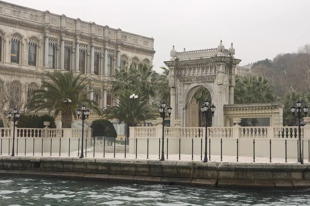 Boğaz'dan Çırağan Sarayı; mermer cephe, yanında oymalı taş kapı, palmiyeler ve kıyıdaki beyaz korkuluk.