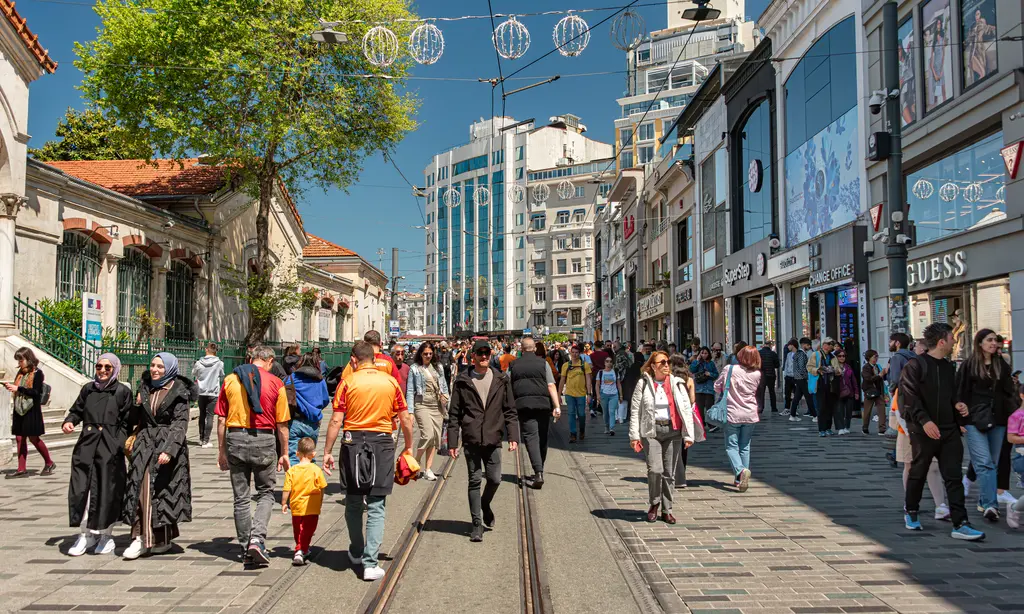 Güneşli bir günde İstiklal Caddesi; ortadaki tramvay raylarında yürüyen insanlar, solda kiremit çatılı alçak yapılar ve sağda mağazalar.