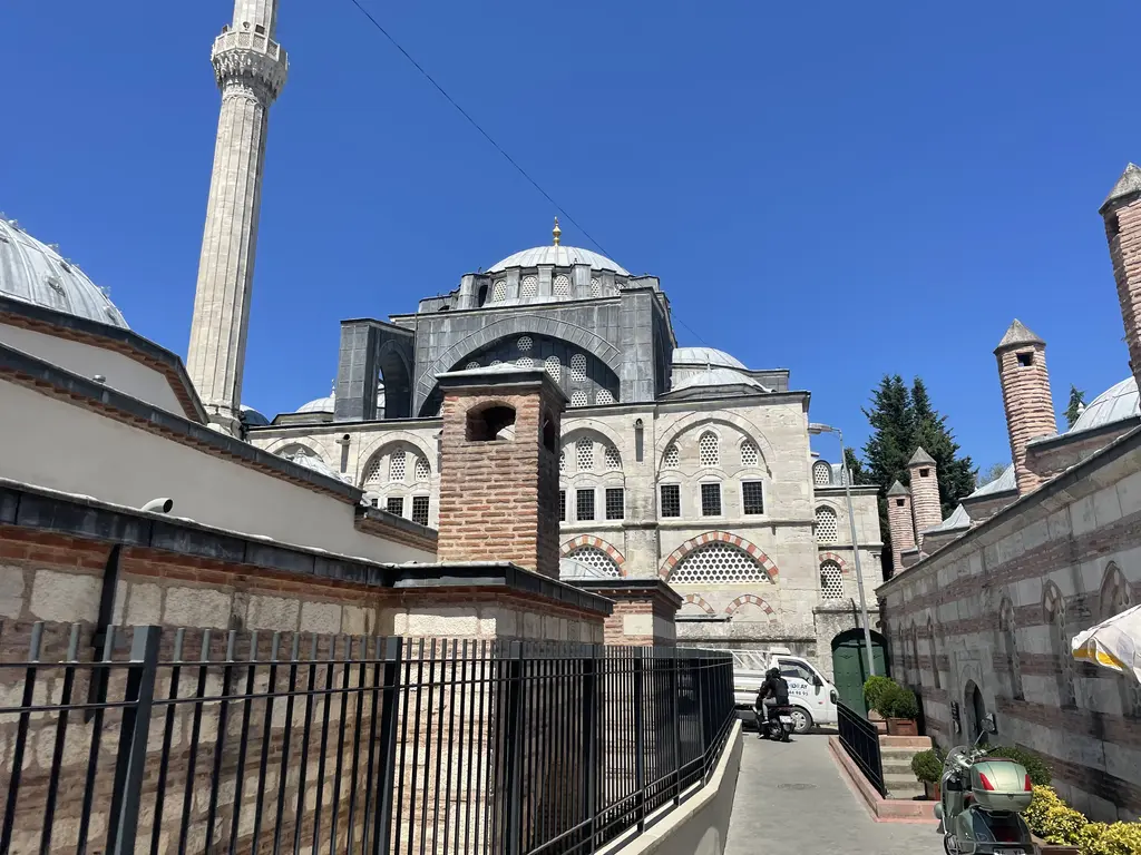Kılıç Ali Paşa Külliyesi'nde dar bir sokak; solda hamamın tuğla bacaları ve demir parmaklık, arkada caminin kubbeleri ve minaresi.