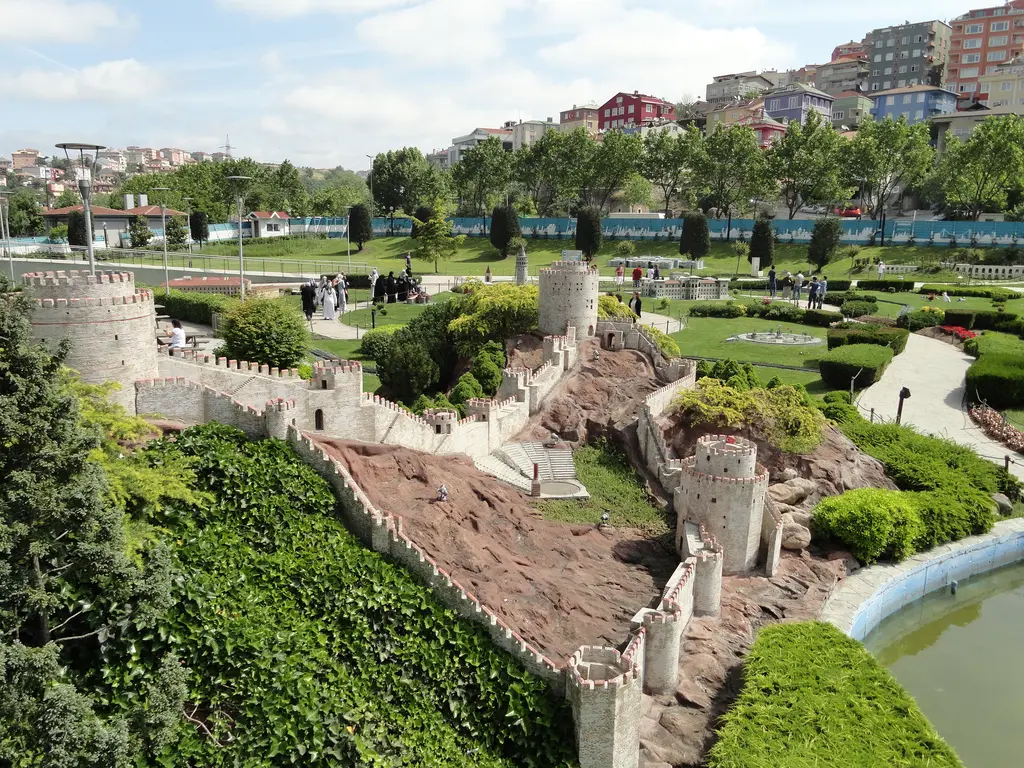 Miniatürk'te Rumeli Hisarı maketi; yamaçtan aşağı inen surlar ve kuleler, kızıl kayalık zemin, sarmaşıklar ve sağ altta havuz kenarı.