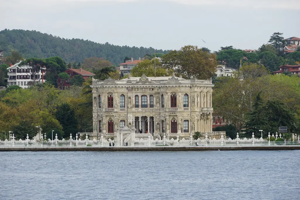 Denizden Küçüksu Kasrı; Boğaz kıyısında beyaz kasır, önünde parmaklıklı rıhtım, arkada ormanlık yamaç ve yalılar.