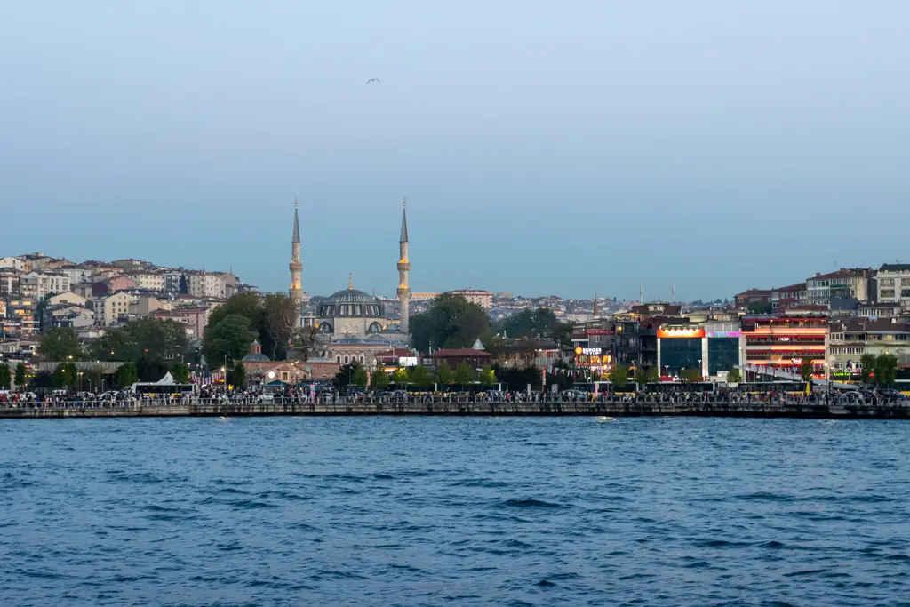 Denizden Üsküdar; kıyı boyunca kalabalık sahil, ortada iki minareli büyük cami ve yamaçta sıralanan binalar.