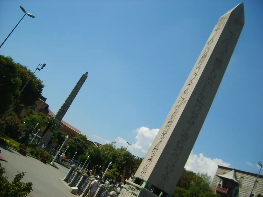 Eğik açıdan Sultanahmet Meydanı; önde Dikilitaş'ın granit gövdesi, arkada Örme Sütun, ağaçlar, sokak lambası ve mavi gök.