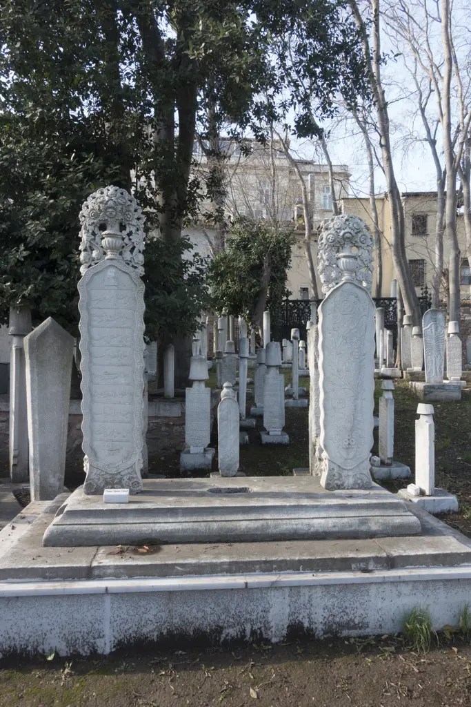 Hazirede tepesi sikke biçiminde oyulmuş uzun mermer mezar taşı; çevresinde başka taşlar ve ağaçlar.