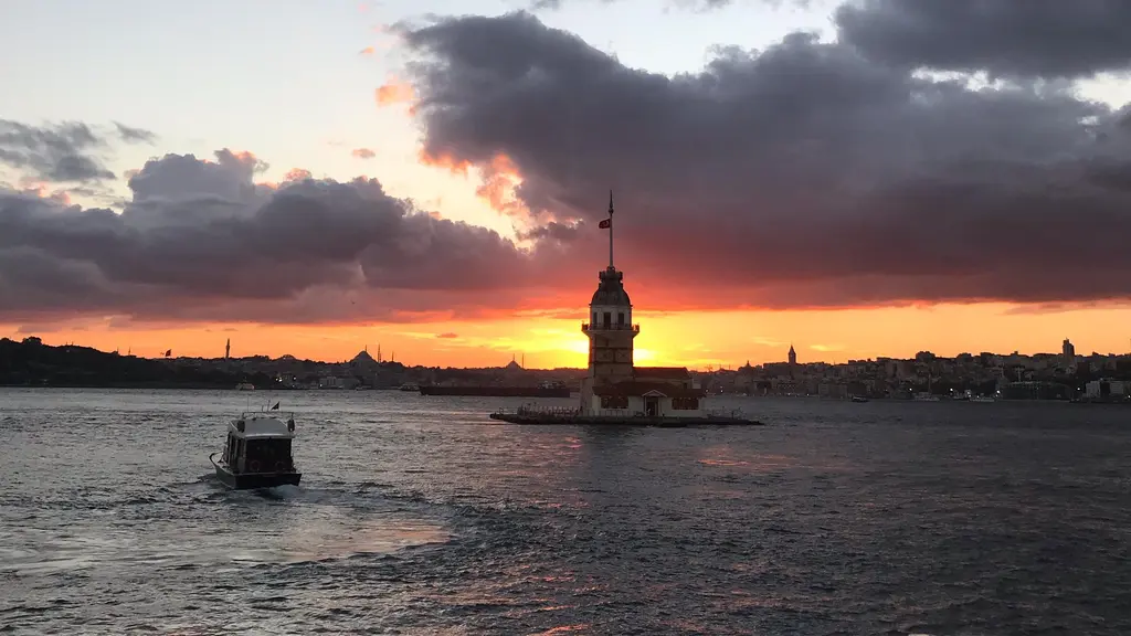 Gün batımında Kız Kulesi; koyu bulutların arasından kızıl ışık, denizde küçük bir tekne ve uzakta kule.