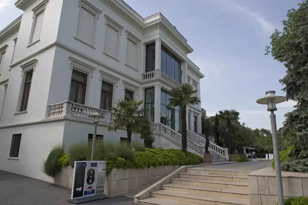 Atlı Köşk'ün köşe cephesi; beyaz duvar, kemerli pencereler, balkonlar, merdiven ve önünde sergi panosu.