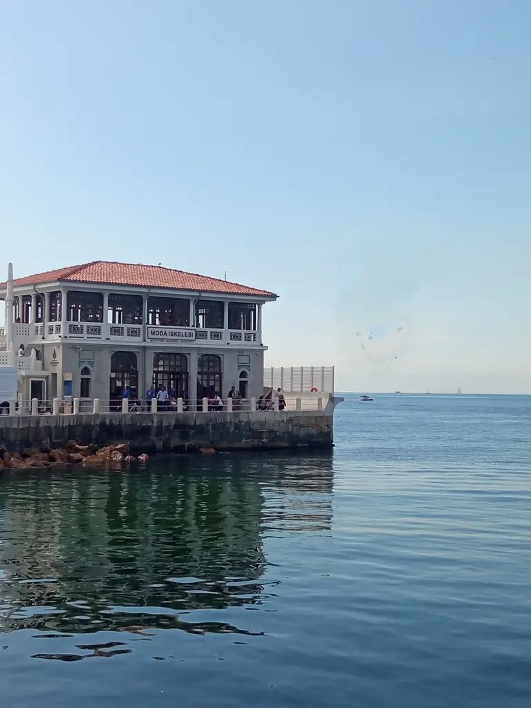 Moda İskelesi; sakin deniz yüzeyinde yansıyan bina, kiremit çatı, beyaz korkuluklar ve açık mavi gök.