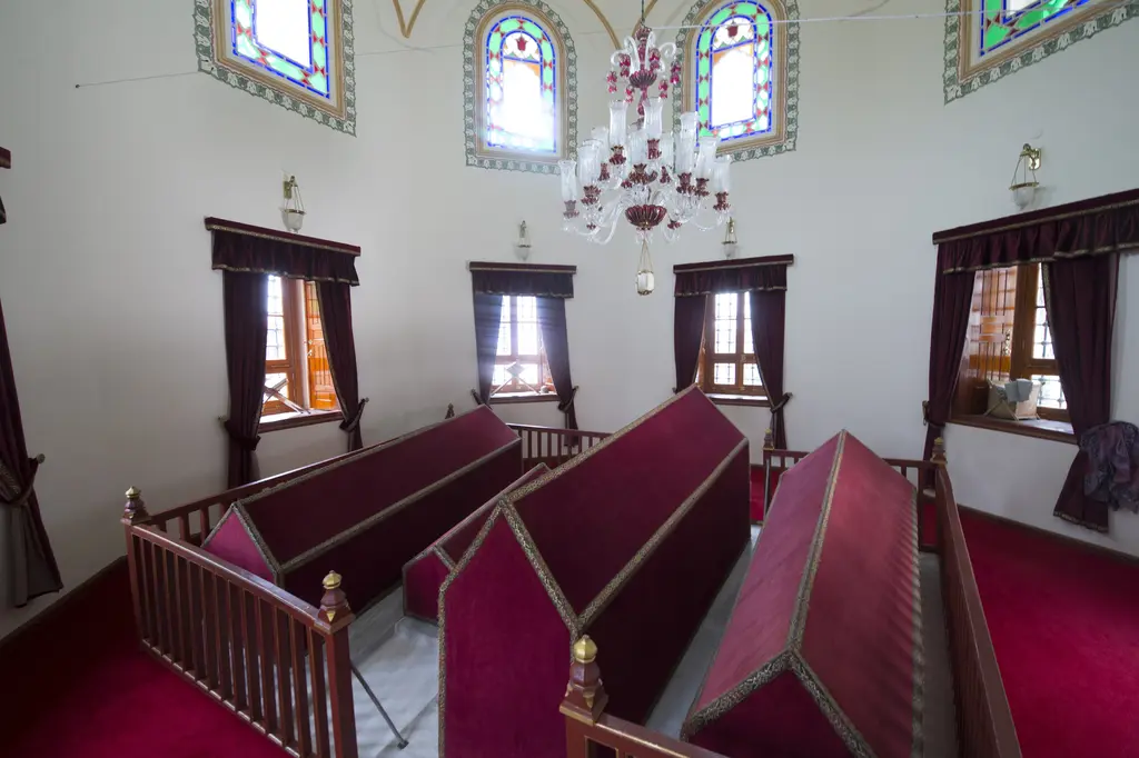 Fatih'te Gülbahar Hatun Türbesi'nin içi; badanalı beyaz duvarlar, mavi yeşil tavan bordürü, renkli camlı kemerli pencereler ve alçak ahşap korkuluk ardında bordo örtülü sandukalar.