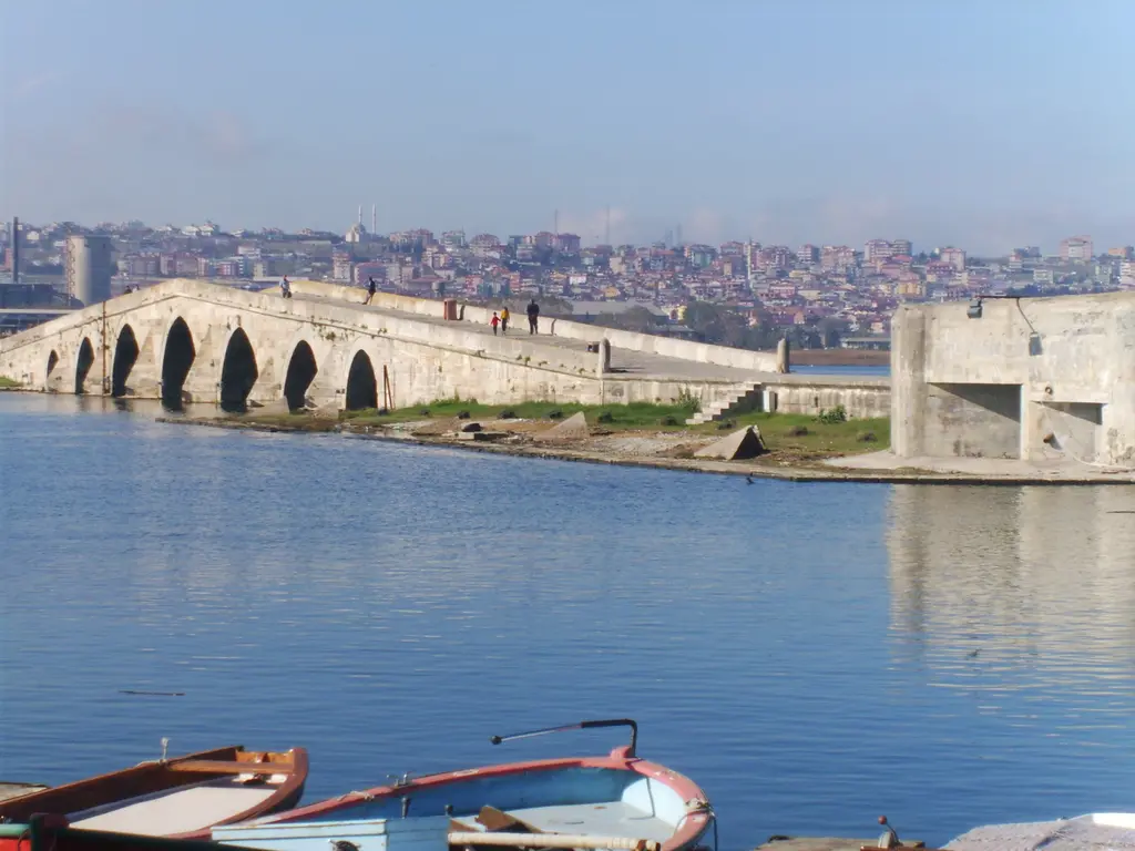 Gölden Kanuni Sultan Süleyman Köprüsü; sakin suda kayıklar, uzanan kemer dizisi ve arkada şehir.