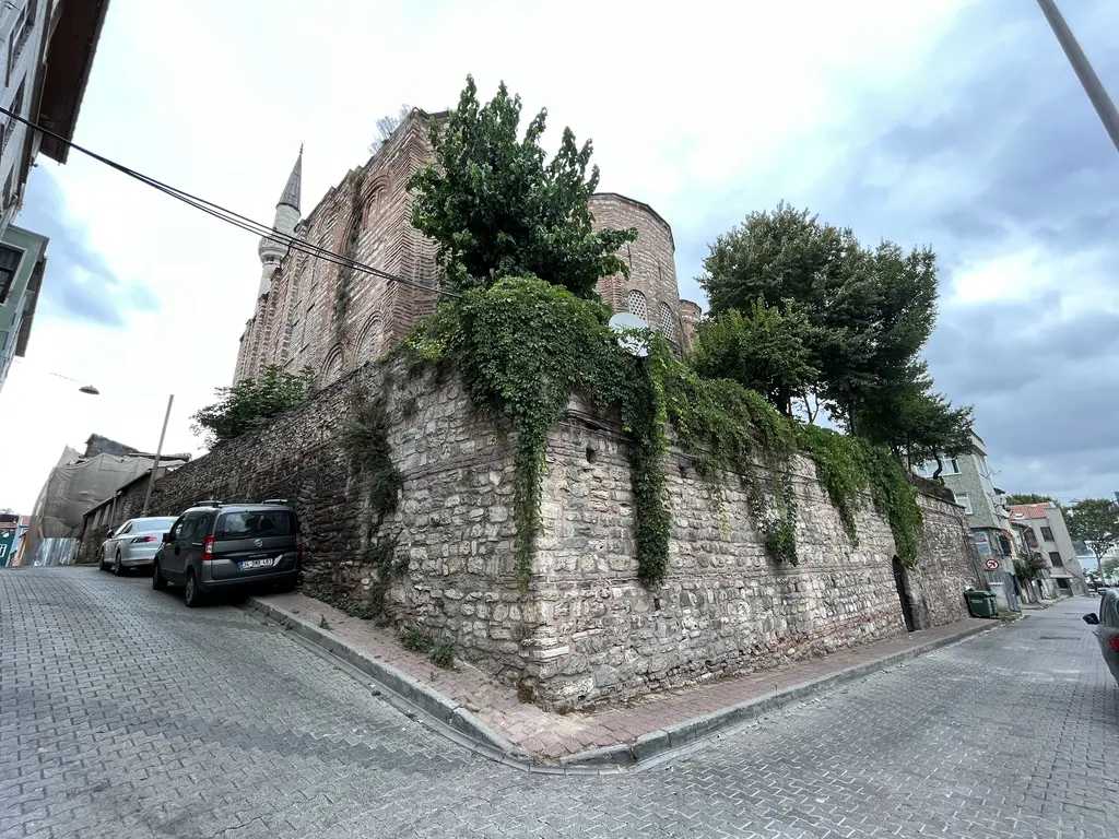 Gül Camii'ne yokuştan bakış; sarmaşıklı yüksek istinat duvarı, tuğla gövde, minare, arnavut kaldırımı ve park etmiş arabalar.