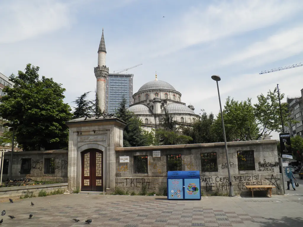 Şişli Meydanı'nda Şişli Camii; taş avlu duvarı ve ahşap kanatlı kapı, arkada kurşun kaplı kubbeler, ince minare ve bir gökdelen.