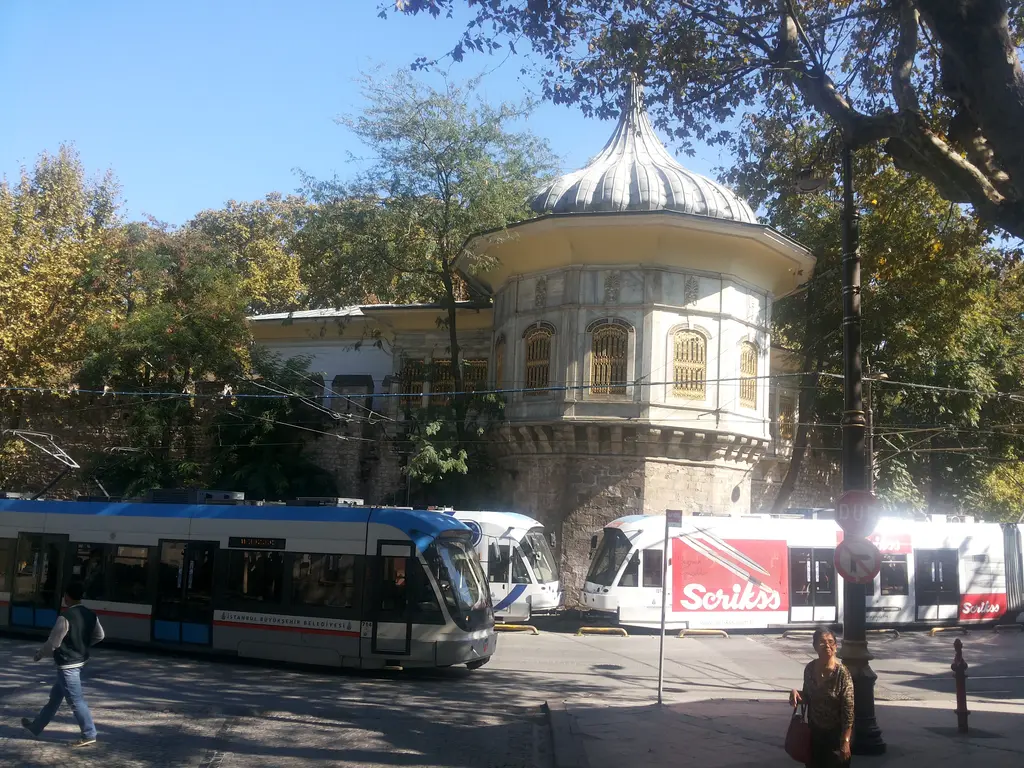 Alay Köşkü önünde duran tramvaylar; ağaçlar arasında soğan kubbeli köşk, burç, yandaki kanat ve güneşli gök.