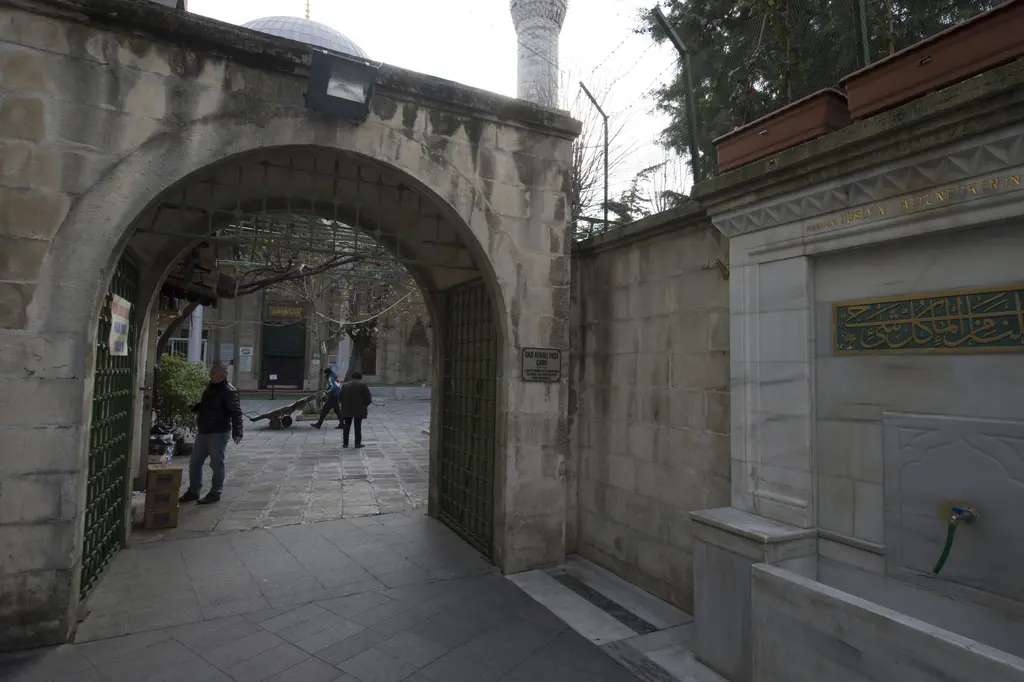 Gazi Atik Ali Paşa Camii'nin avlu kapısı; taş kemerli giriş, yeşil demir kanat, içeride avluda yürüyen insanlar ve sağda yazılı çeşme duvarı.