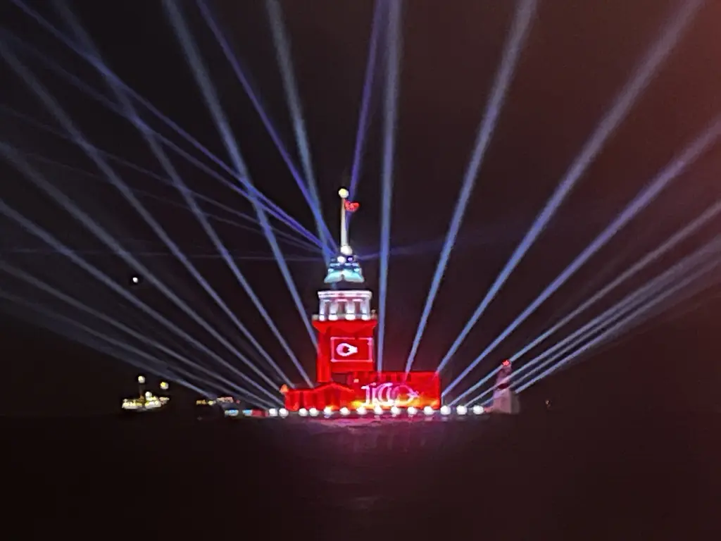 Gece Kız Kulesi; kırmızı ışıklarla aydınlatılmış kule, çevresinden göğe yükselen lazer ışınları ve karanlık deniz.