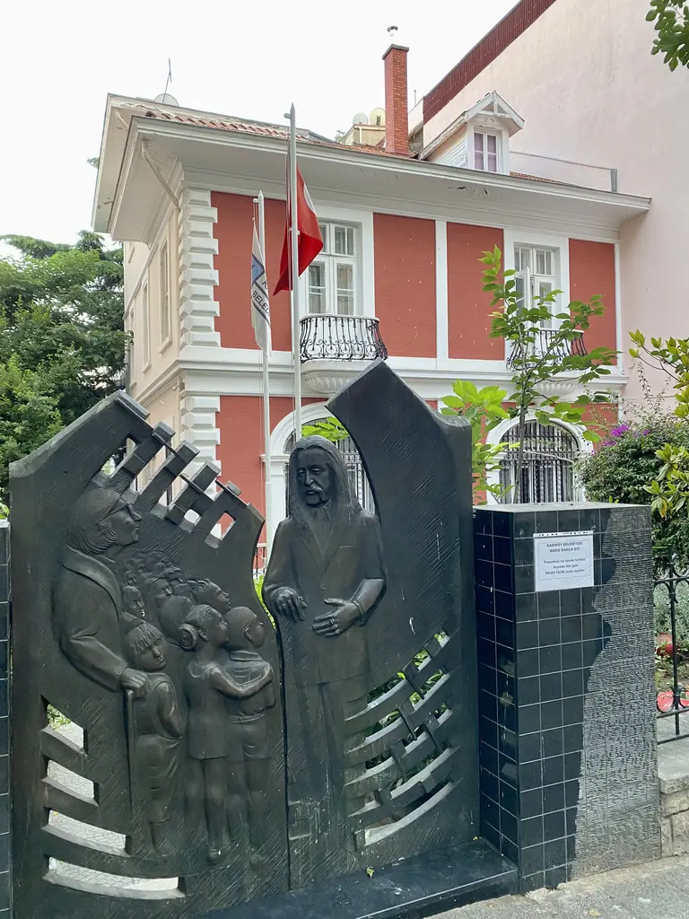 Barış Manço'nun evi; kırmızı ve beyaz cepheli köşk, Türk bayrağı, önde siyah kabartmalı anıt ve ağaçlar.