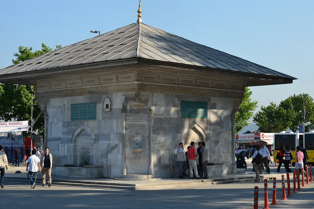 III. Ahmed Çeşmesi; mavi gökyüzü altında kare gövde, yeşil kitabeler, köşe musluğu ve önde sarı taksi.