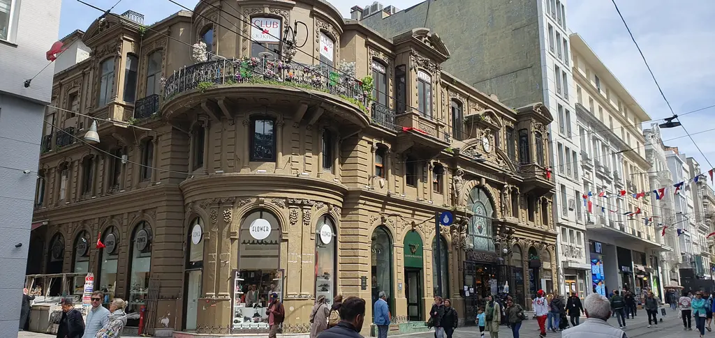 İstiklal Caddesi'nde Çiçek Pasajı; kahverengi taş cephe, yuvarlak köşe, demir balkonlar, sağda caddeye uzanan binalar ve kalabalık.