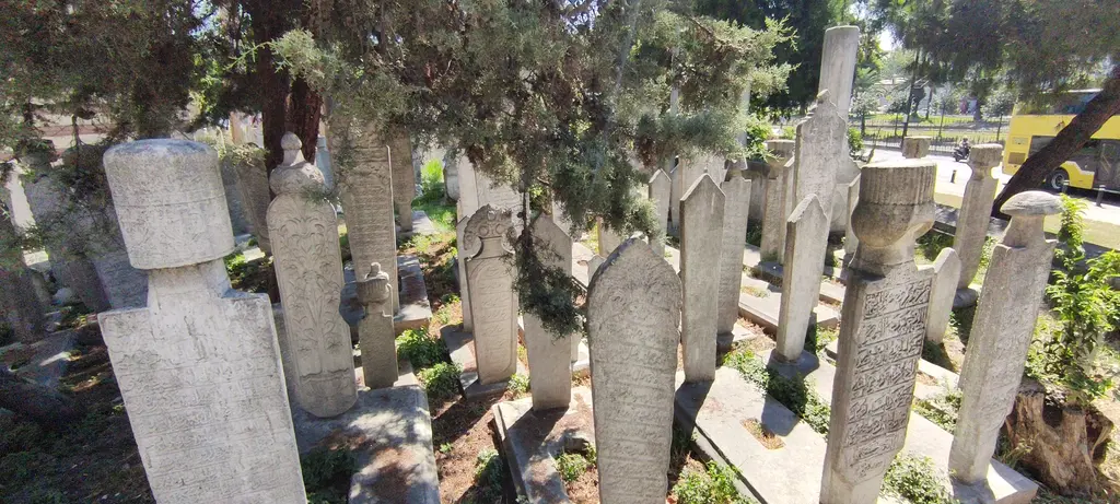 Murat Paşa Camii hazire; ağaç gölgesinde sıralı mermer mezar taşları, sarıklı ve yazıtlı taşlar ve arkada avlu duvarı.