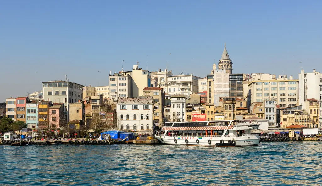 Haliç'ten Karaköy kıyısı; rıhtım boyunca renkli binalar, iskelede beyaz bir yolcu teknesi ve yamacın üstünde Galata Kulesi.
