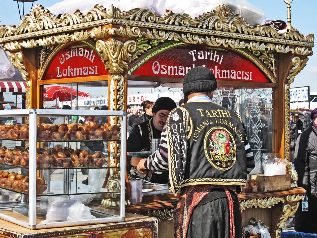 Eminönü meydanında lokma tezgâhı; yaldızlı süslü araba, tepsilerde lokmalar ve geleneksel kıyafetli satıcı.