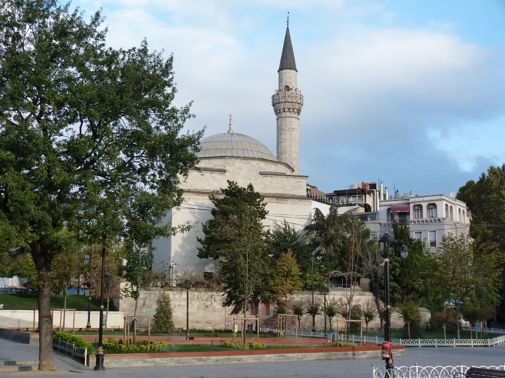Firuz Ağa Camii; yeşil ağaçlar arasında kubbe ve minare, önde çiçek tarhı, meydan ve arka planda binalar.