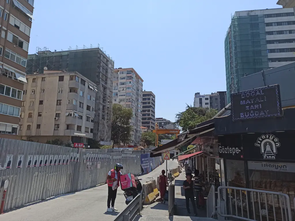 Göztepe'de istasyon çevresi; inşaat panoları, büfe ve tabelalar, kuryeler ve iki yanda yüksek apartmanlar.