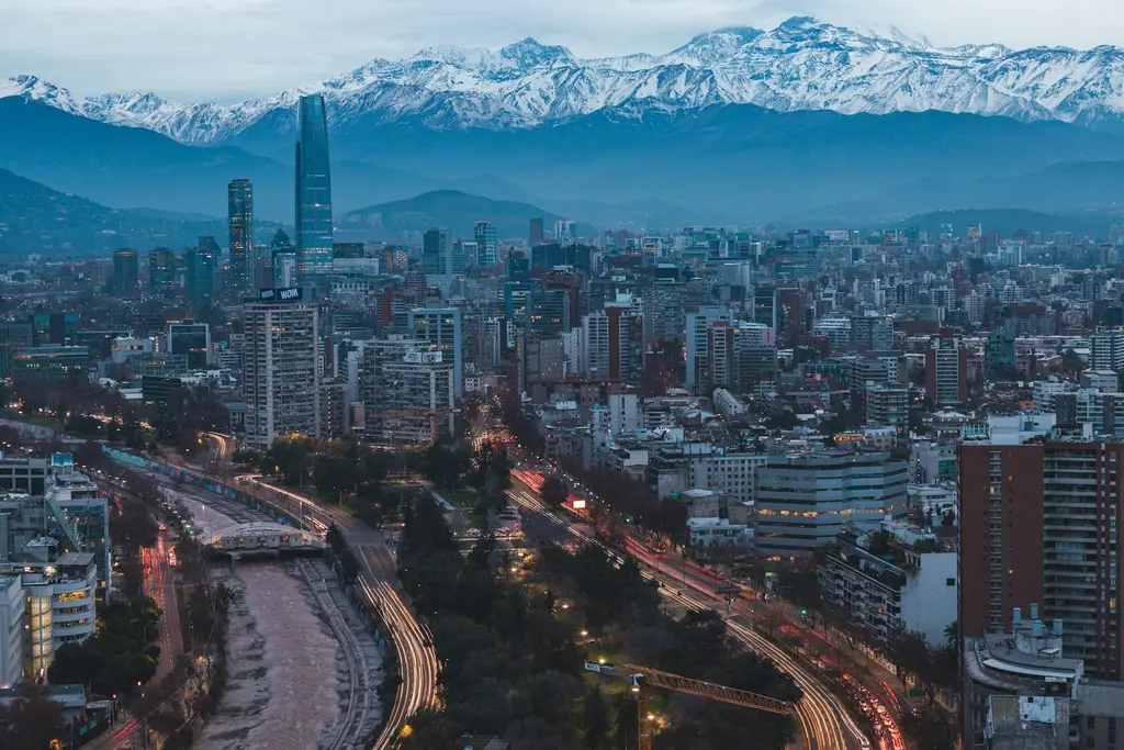 Şili'nin başkenti Santiago'nun alacakaranlıkta çekilmiş silüeti; şehri kesen nehir ve trafik ışıkları, gökdelenlerin arkasında karlı And Dağları.