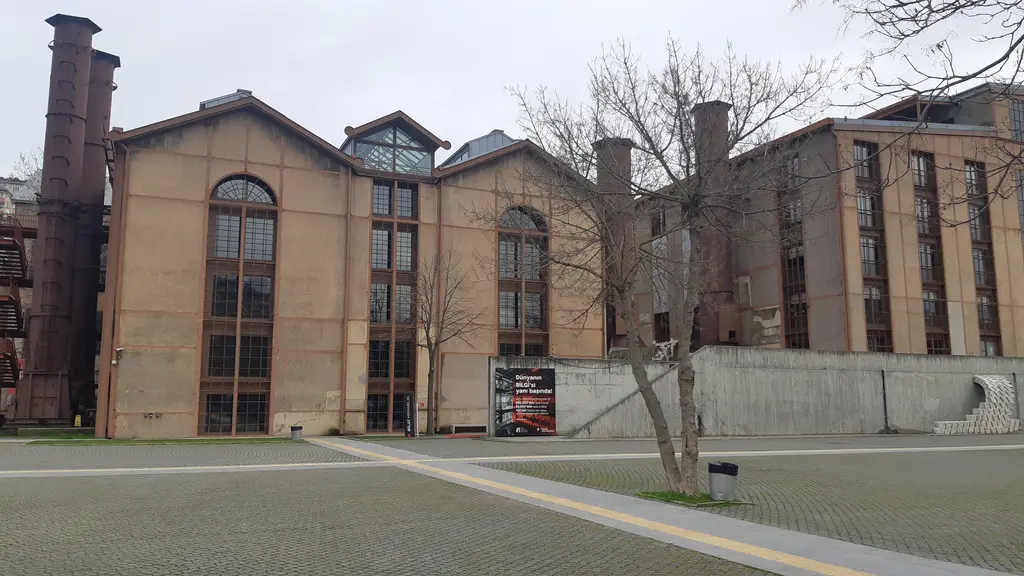 Santralistanbul'da makine dairesi; kemerli büyük pencereli tuğla cephe, yanında iki demir baca, önde taş meydan ve ağaç.