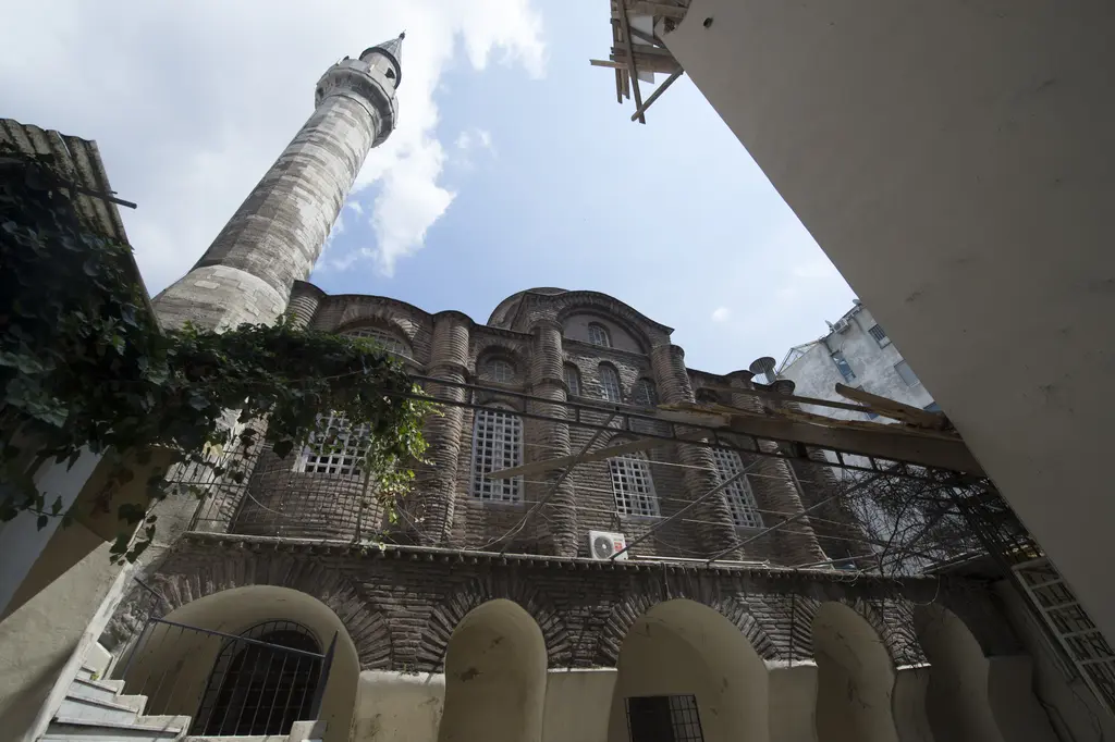 Bodrum Camii'ne avludan dik alttan bakış; altta alt yapının beyaz sıvalı kemer dizisi, üstte tuğla kilise gövdesi ve payandalar, solda minare ile sarmaşık.
