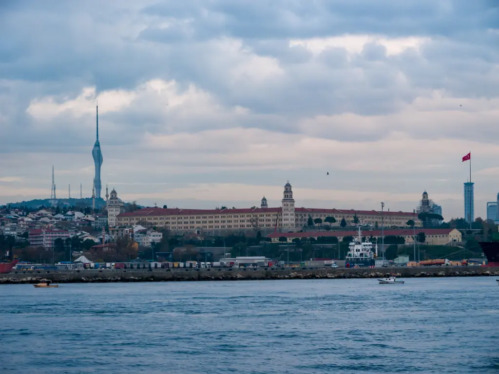 Denizden Üsküdar; kıyıda uzun kışla binası, arkasında yamaçtaki evler ve tepede yükselen ince Çamlıca Kulesi.