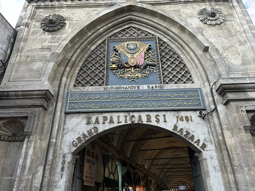 Kapalıçarşı'nın Nuruosmaniye Kapısı; sivri taş kemer, üstünde altın renkli tuğra ile çarşının adını taşıyan levhalar, iki Türk bayrağı ve önünde kalabalık.